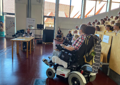 Rollstuhlfahrer im Vordergrund. Weiter Gäste hören einem Referenten zu.